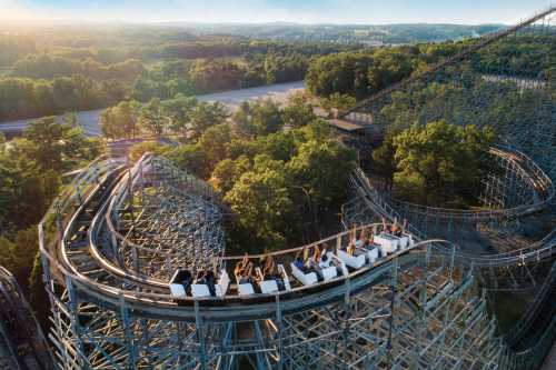 Aerial view of a wooden roller coaster with riders enjoying the thrill, surrounded by lush green trees and hills.