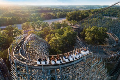 Aerial view of a wooden roller coaster with riders enjoying the thrill, surrounded by lush green trees and hills.