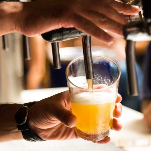 A hand pours a golden beer into a glass from a tap, with blurred figures in the background.