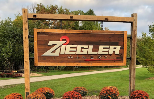 Sign for Ziegler Winery, featuring a wooden frame and colorful flowers in front, set against a cloudy sky.