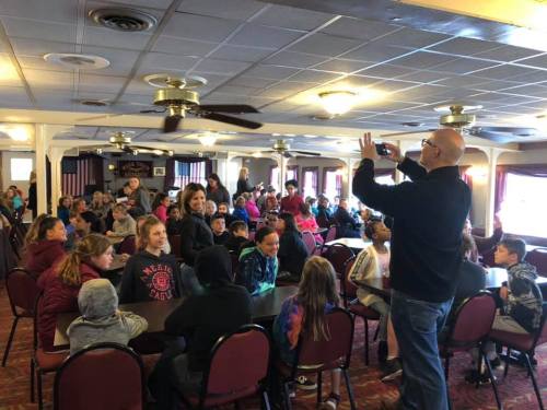 A large group of children and adults gathered in a room, with one person taking a photo of the audience.