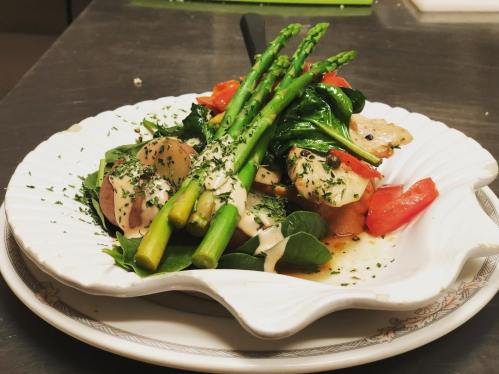A plated dish featuring grilled chicken, asparagus, spinach, tomatoes, and a drizzle of sauce, garnished with herbs.