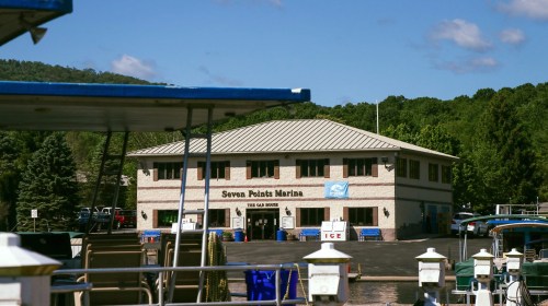 A marina building with "Seven Points Marina" sign, surrounded by trees and blue sky, with boats in the foreground.