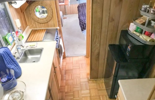 A cozy kitchen with wooden walls, a sink, stove, and a view into a bedroom area with carpeted flooring.