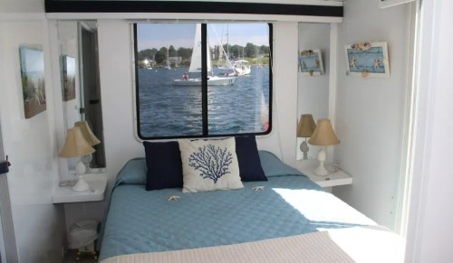 Cozy bedroom with a blue quilt, lamps, and a view of boats on the water through a large window.