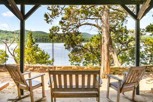 A serene lakeside view from a porch with two wooden chairs, surrounded by trees and mountains in the background.