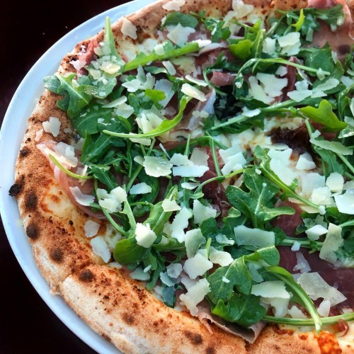 A close-up of a pizza topped with arugula, prosciutto, and shaved cheese on a golden crust.
