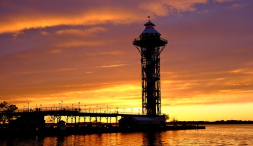 Silhouette of a tall tower against a vibrant sunset over a calm body of water.