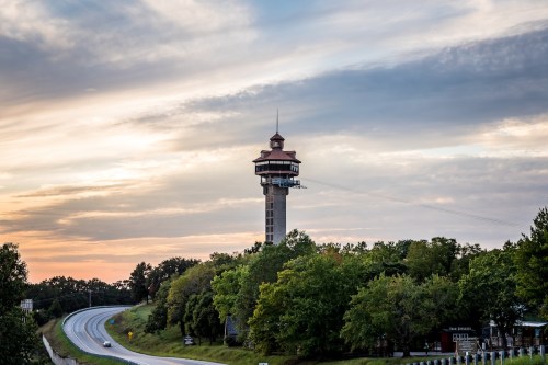 A tall observation tower stands near a highway, surrounded by trees under a colorful sunset sky.