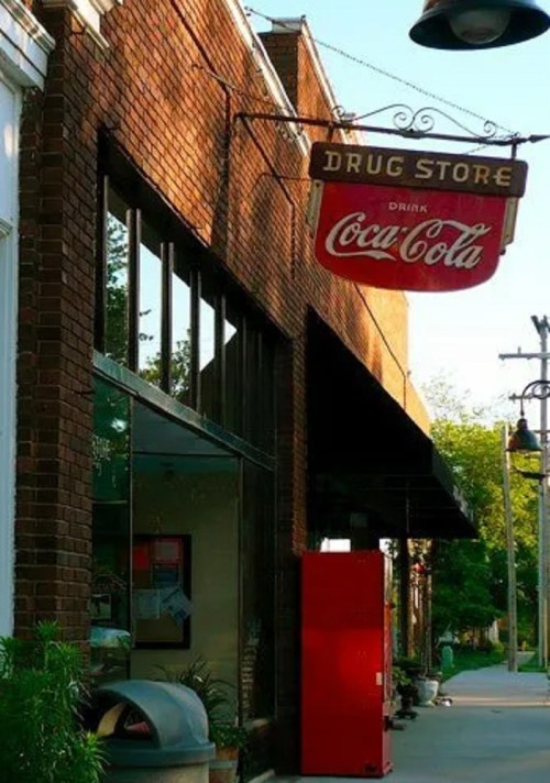 A vintage drug store with a Coca-Cola sign, featuring brick architecture and a red vending machine outside.