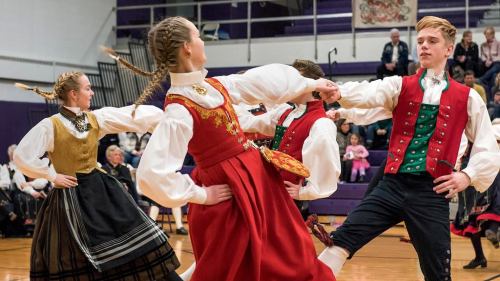 A group of dancers in traditional costumes perform a lively folk dance on a gymnasium floor.