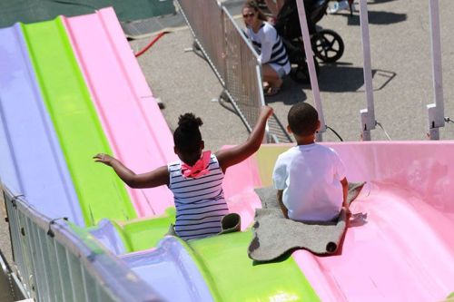Two children slide down a colorful slide at a fair, with a parent watching in the background.