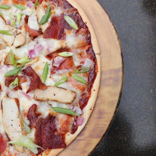 A close-up of a pizza topped with chicken, bacon, onions, and green onions on a wooden serving board.
