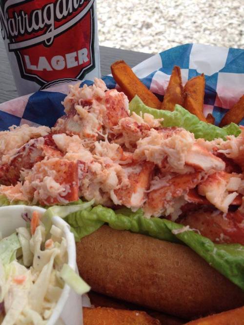 A lobster roll topped with shredded lobster on lettuce, served with sweet potato fries and a beer in the background.
