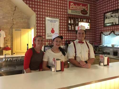 Three staff members in retro diner attire smile behind a counter with vintage decor and condiments.