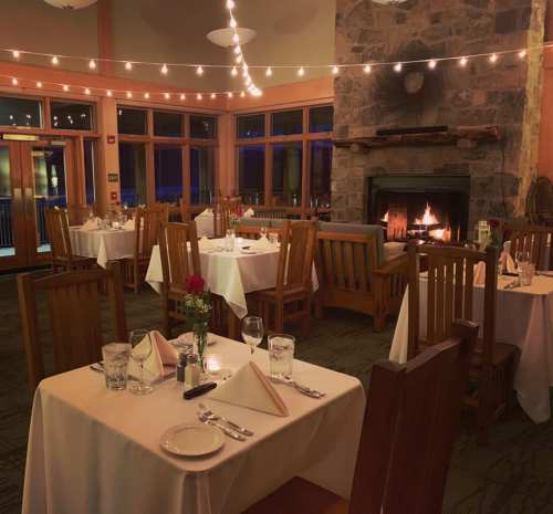 Cozy restaurant interior with tables set for dining, warm lighting, and a fireplace in the background.