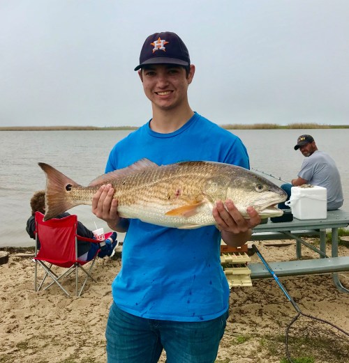 A young man in a blue shirt and cap holds a large fish by a lake, with people sitting in the background.