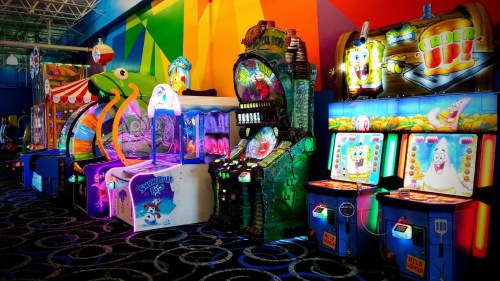 Colorful arcade games line a vibrant wall, featuring various themed machines and bright lights.