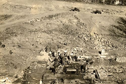 Historical black-and-white photo of workers at a construction site, with machinery and rocky terrain in the background.