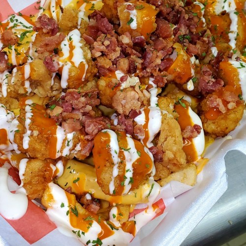 A close-up of loaded fries topped with cheese, bacon bits, drizzled with ranch and spicy sauce, garnished with parsley.
