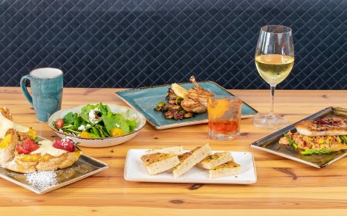 A table spread with various dishes: salad, grilled chicken, toast, and drinks, including a glass of white wine.