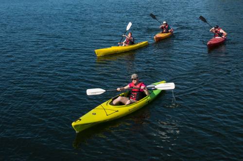 Four people kayaking on a calm blue lake, wearing life jackets in yellow, green, and red kayaks.