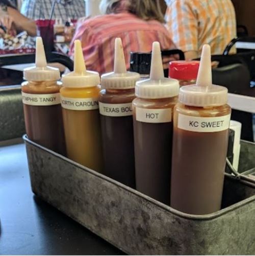 A metal caddy holds six bottles of barbecue sauces labeled Memphis Tangy, North Carolina, Texas Bold, Hot, and KC Sweet.