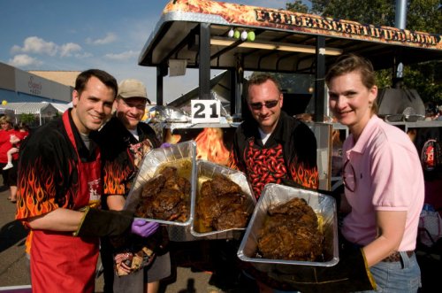Four people in flame-patterned shirts hold trays of cooked meat in front of a food truck at an outdoor event.