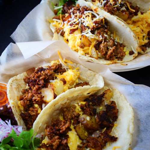 Two tacos filled with seasoned meat, potatoes, and cheese, served with fresh toppings and a side of salsa.