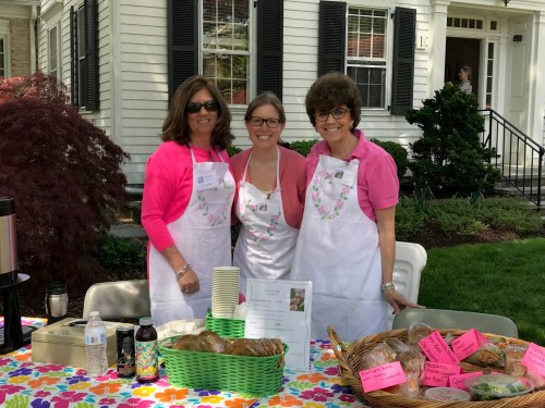 Three women in pink shirts and white aprons smile behind a table with baked goods and drinks at a community event.