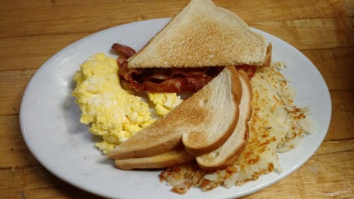 A plate with scrambled eggs, crispy bacon sandwich, and golden brown hash browns on a wooden table.