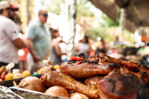 Close-up of grilled chicken legs on a tray, with people mingling in the background at a lively outdoor event.