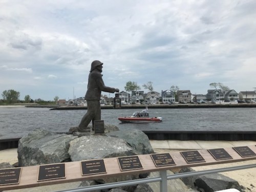 A statue of a fisherman holding a lantern stands by the water, with houses and a boat in the background.