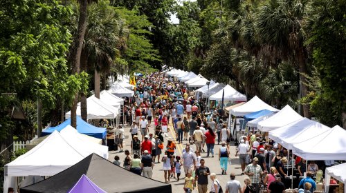A bustling street fair with numerous tents and a large crowd of people enjoying the event under palm trees.
