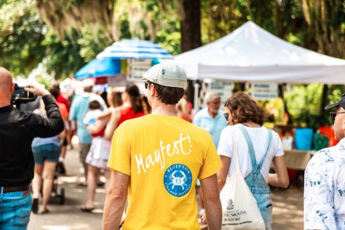 A person in a yellow shirt walks through a lively outdoor market, surrounded by tents and people enjoying the festival.