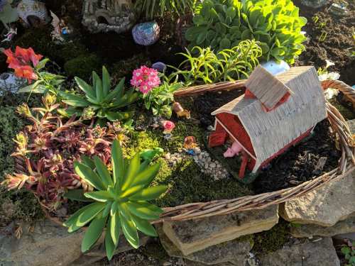 A miniature garden scene featuring a small red barn, colorful flowers, and decorative plants in a woven basket.