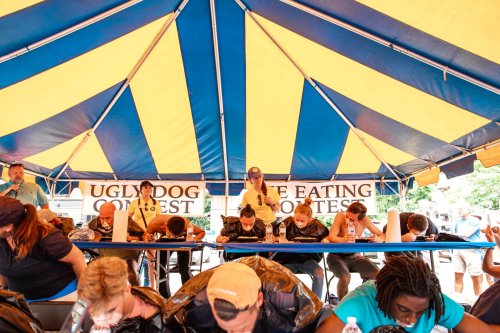 Participants compete in a hot dog eating contest under a colorful tent, with spectators watching in the background.