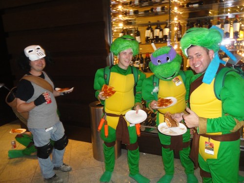 Four people in Teenage Mutant Ninja Turtles costumes hold plates of food, while one person in a mask stands nearby.