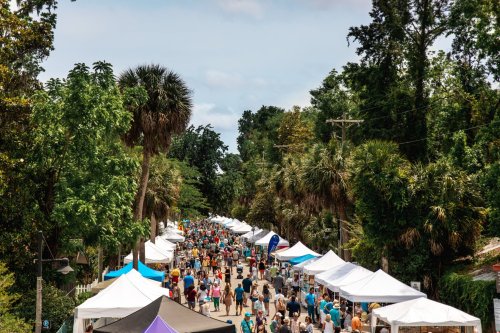 A bustling outdoor market with numerous tents and a crowd of people among lush greenery and palm trees.