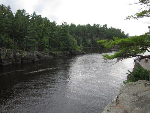 A serene river flows through a lush green landscape, bordered by rocky banks and trees under a cloudy sky.
