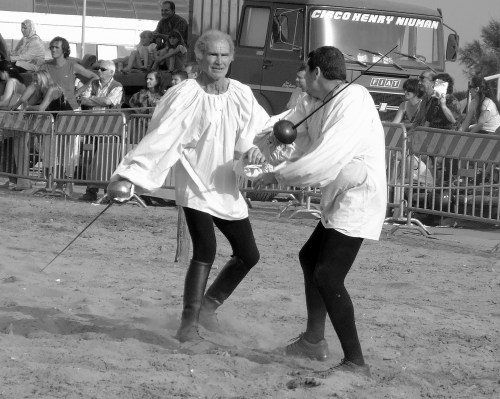 Two men in historical costumes engage in a sword fight on a sandy beach, with a crowd and a circus truck in the background.