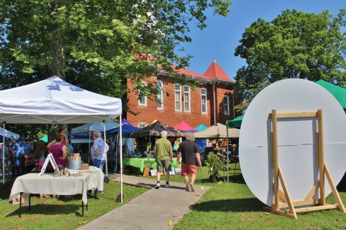 A vibrant outdoor market scene with tents, people walking, and a historic building in the background under a clear blue sky.
