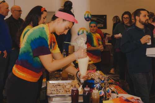 A person in a tie-dye shirt decorates desserts at a festive event, surrounded by a lively crowd.