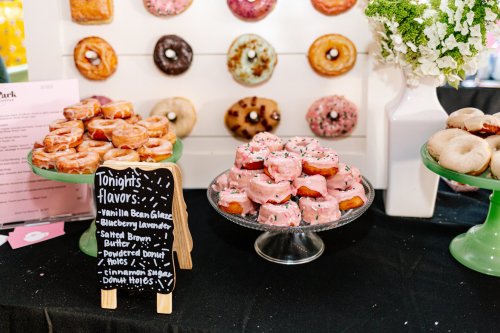 A display of colorful donuts on stands, with a chalkboard listing flavors like vanilla bean glaze and blueberry lavender.