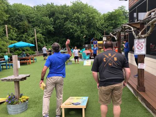 Two men play cornhole outdoors in a vibrant, green space with people enjoying the area in the background.