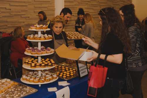 A woman serves donuts from a tiered display while customers select treats at a dessert event.