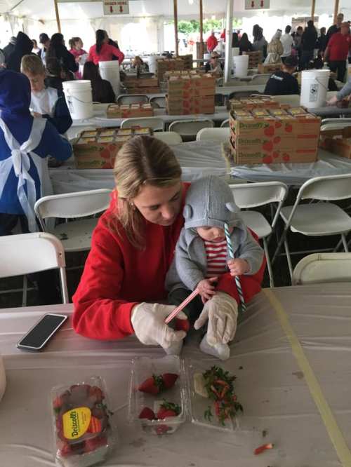 A woman in a red hoodie helps a child with a straw while sitting at a table with strawberries in a tented area.