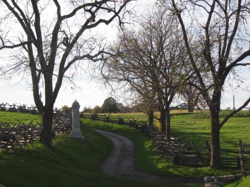 A winding dirt path leads through a grassy area with bare trees and a stone monument, surrounded by a wooden fence.