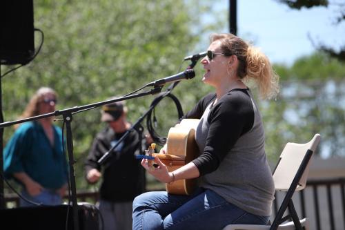 A woman sings and plays guitar on stage, while two people are visible in the background enjoying the performance.