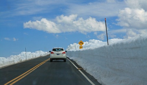 A car drives on a snowy road with high snowbanks and a warning sign indicating a curve ahead.
